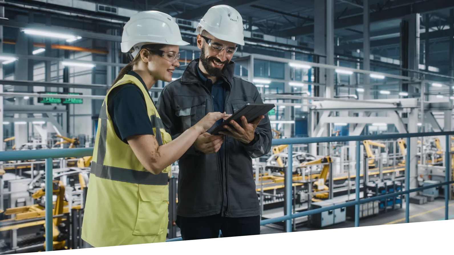 A man and a woman dressed in high visibility vests, using a tablet. It looks like they are in a manufacturing facility.
